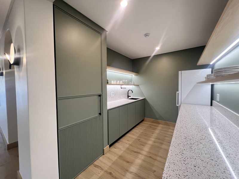 Modern office kitchenette featuring sage green cabinetry, terrazzo countertops, floating shelves with integrated LED lighting, and high-quality wood-look flooring.