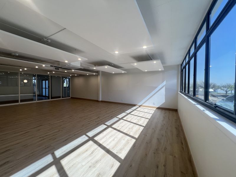 A bright, open-plan office space featuring modern wood-look flooring, suspended acoustic ceiling panels, and large windows providing ample natural light.