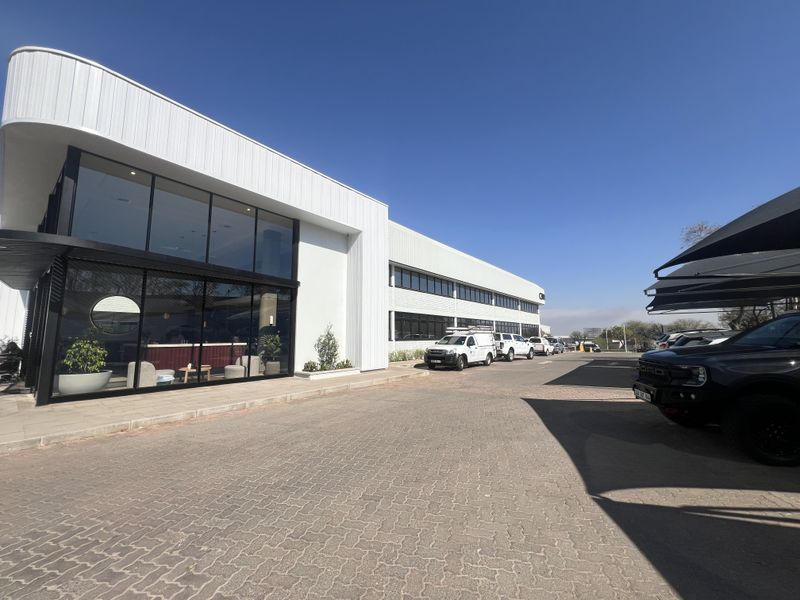 Modern office building exterior featuring a prominent glass facade, paved parking area, and covered parking bays under a clear blue sky.
