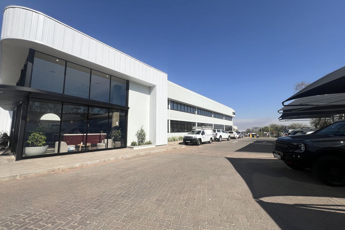 Modern office building exterior featuring a prominent glass facade, paved parking area, and covered parking bays under a clear blue sky.