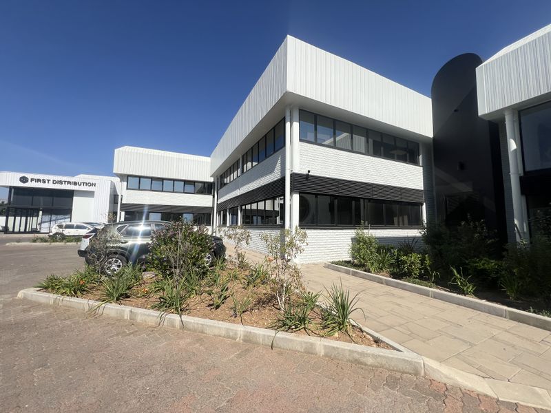 Modern office building exterior featuring white metal cladding, dark aluminum window frames, and a paved pedestrian walkway leading to the entrance.