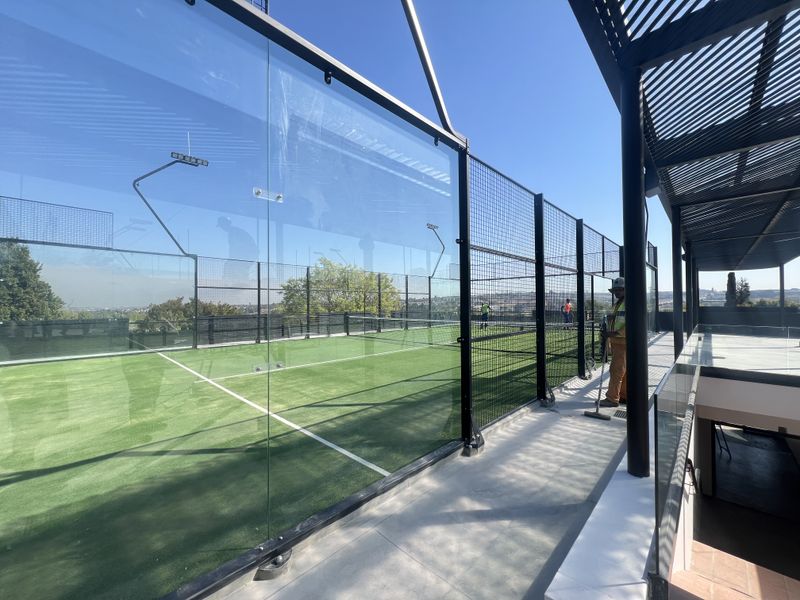 A modern outdoor padel court featuring glass and mesh perimeter fencing, artificial turf surface, and an adjacent covered walkway.