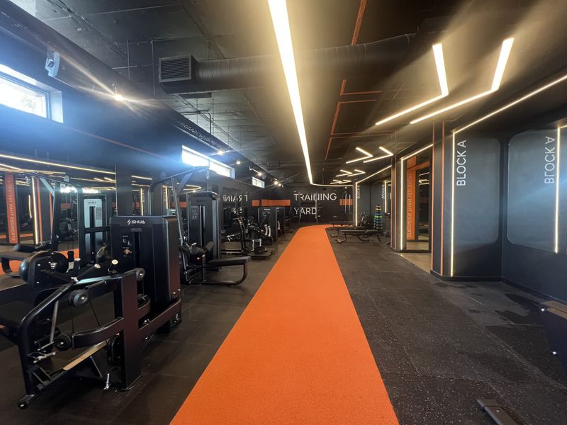A modern, well-equipped commercial gym featuring industrial-style exposed ceilings, linear lighting, mirrored walls, and a dedicated orange running track lane.