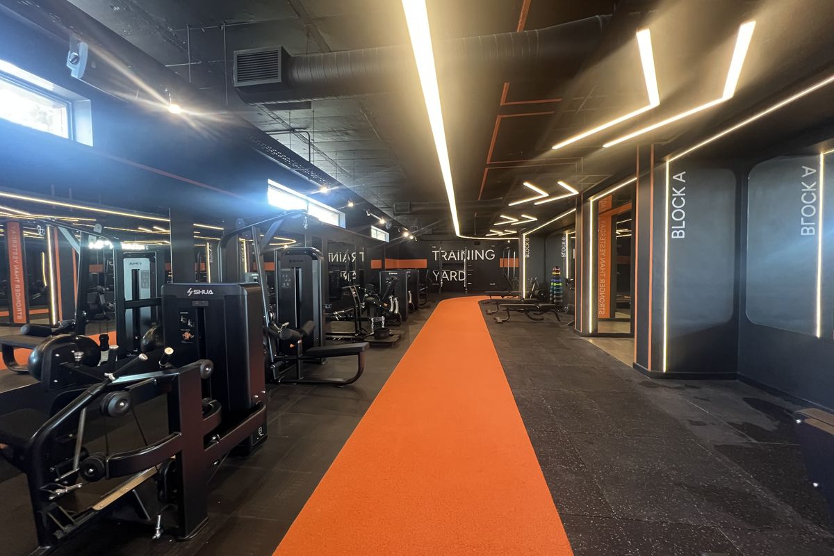 A modern, well-equipped commercial gym featuring industrial-style exposed ceilings, linear lighting, mirrored walls, and a dedicated orange running track lane.