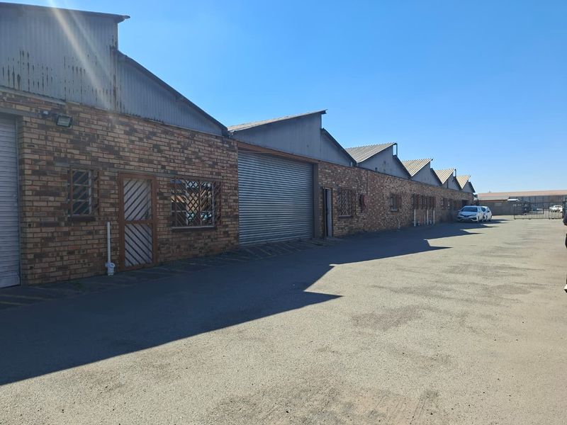 Exterior view of an industrial warehouse complex featuring brick construction, corrugated metal roofing, and multiple roller shutter access doors.