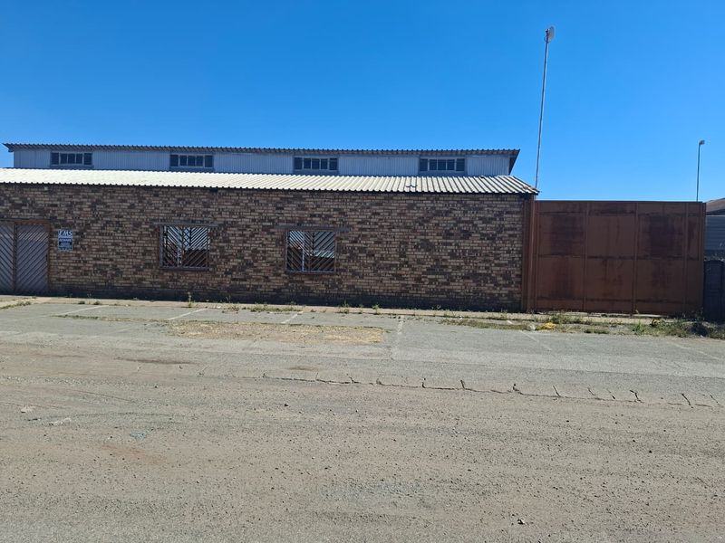 Industrial property exterior featuring a face brick facade, corrugated metal roofing, security-barred windows, and a large metal gate with an adjacent paved parking area.