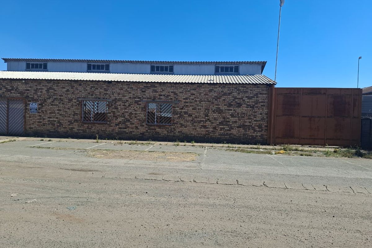 Industrial property exterior featuring a face brick facade, corrugated metal roofing, security-barred windows, and a large metal gate with an adjacent paved parking area.