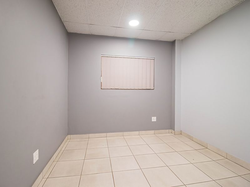 A clean, empty office space featuring neutral grey walls, tiled flooring, and a suspended ceiling with recessed lighting.