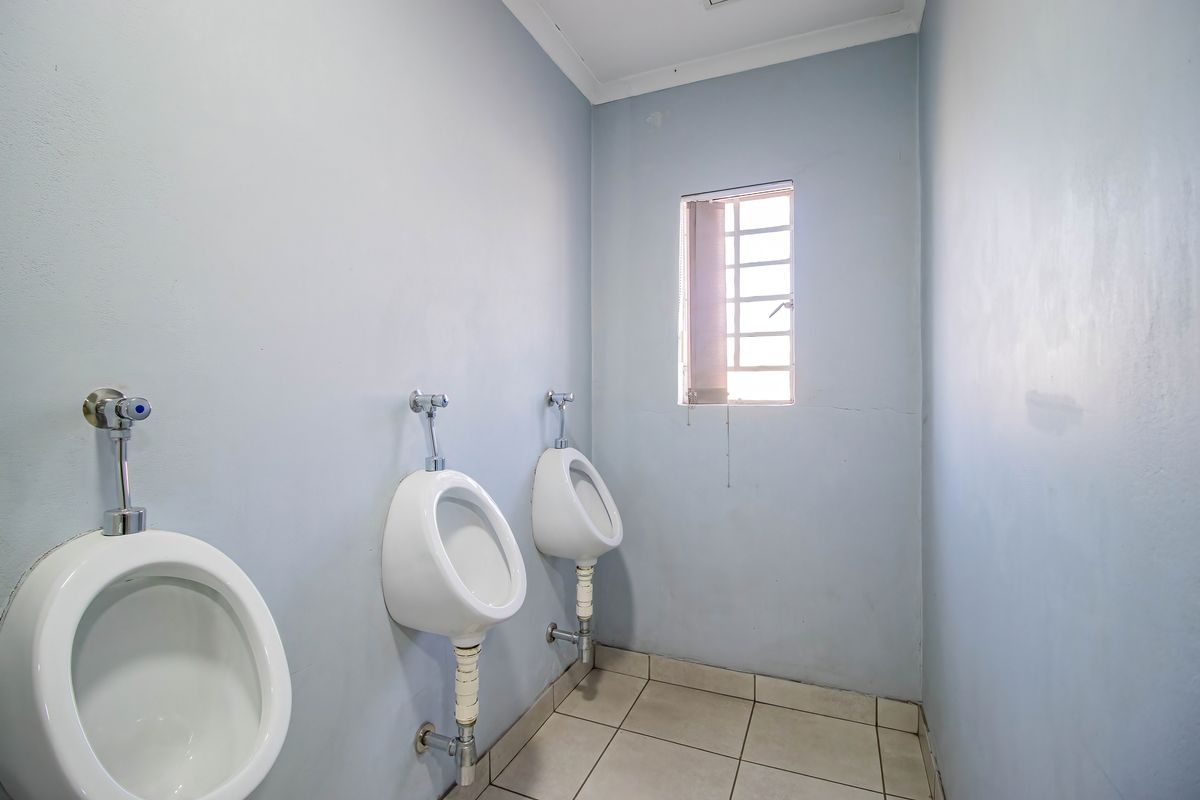 A functional industrial restroom facility featuring multiple wall-mounted urinals, tiled flooring, and a window with security bars.