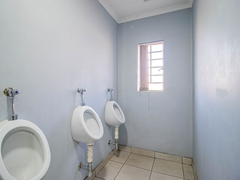A functional industrial restroom facility featuring multiple wall-mounted urinals, tiled flooring, and a window with security bars.