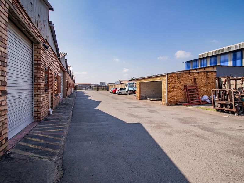 Industrial exterior view featuring a paved driveway, brick warehouse facades with roller shutter doors, and an open-sided storage bay.