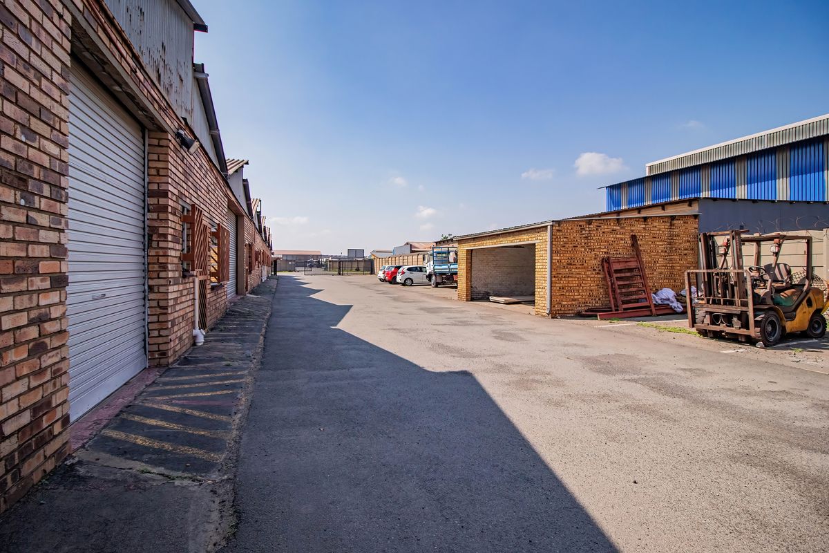 Industrial exterior view featuring a paved driveway, brick warehouse facades with roller shutter doors, and an open-sided storage bay.