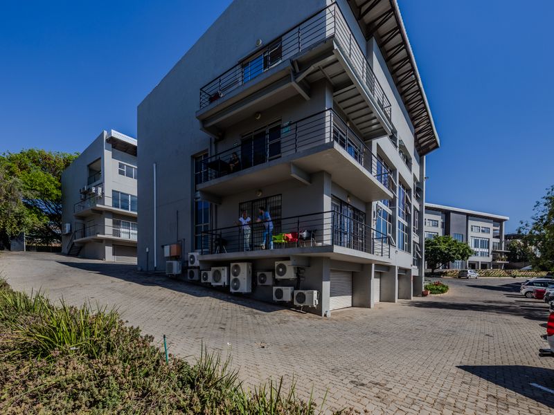 A multi-story office building featuring external balconies, metal railings, and multiple wall-mounted air conditioning units, set within a paved commercial complex.