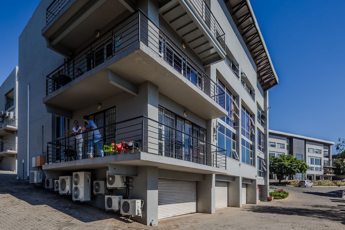 A multi-story office building featuring external balconies, multiple air conditioning units, and ground-level roller shutter garage doors with a paved parking area.