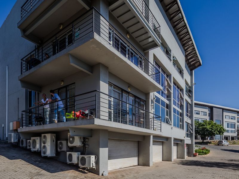 A multi-story office building featuring external balconies, multiple air conditioning units, and ground-level roller shutter garage doors with a paved parking area.
