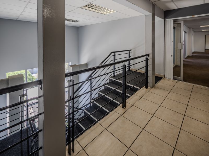 A bright office hallway featuring a staircase with metal balustrades, ceramic tiled flooring, and a suspended ceiling with recessed lighting.