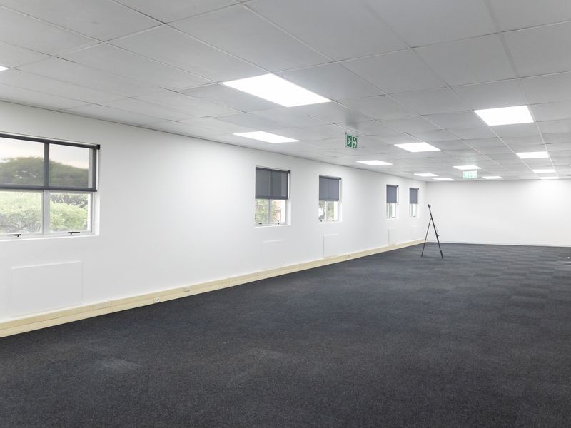 A spacious, open-plan office area featuring neutral white walls, dark carpet tiles, and a suspended ceiling with integrated recessed lighting.