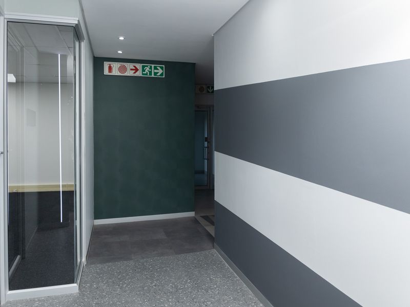 A clean, modern office hallway featuring terrazzo flooring, glass partitioning, and clear emergency exit signage with a contemporary painted wall finish.