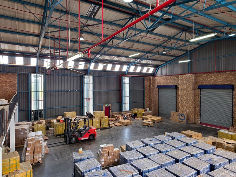 Spacious industrial warehouse featuring high ceilings with exposed steel trusses, brick walls, roller shutter doors, and a mezzanine level for storage or office use.