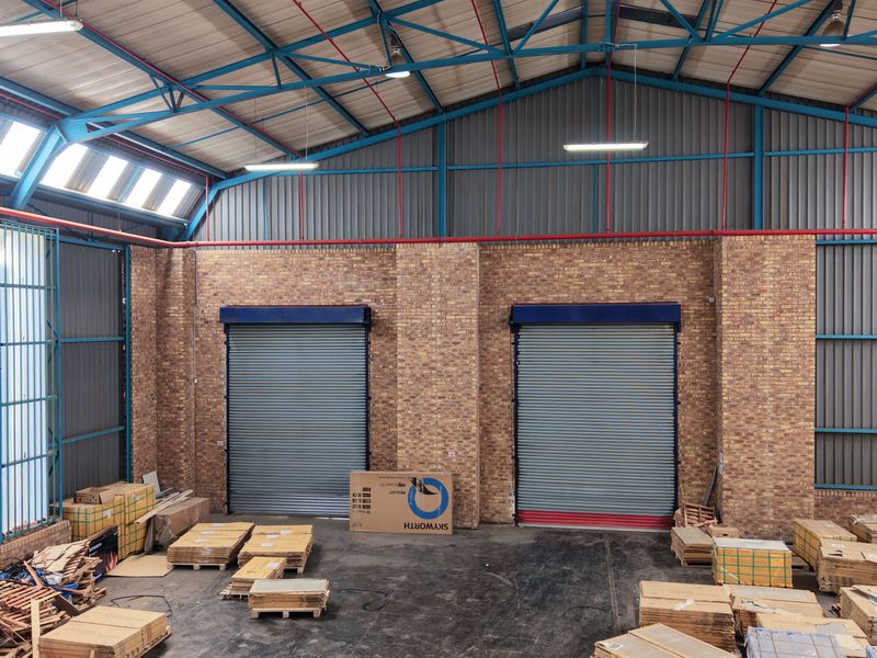 Spacious industrial warehouse unit featuring high ceilings with exposed steel trusses, brick walls, and two large roller shutter doors for easy access.