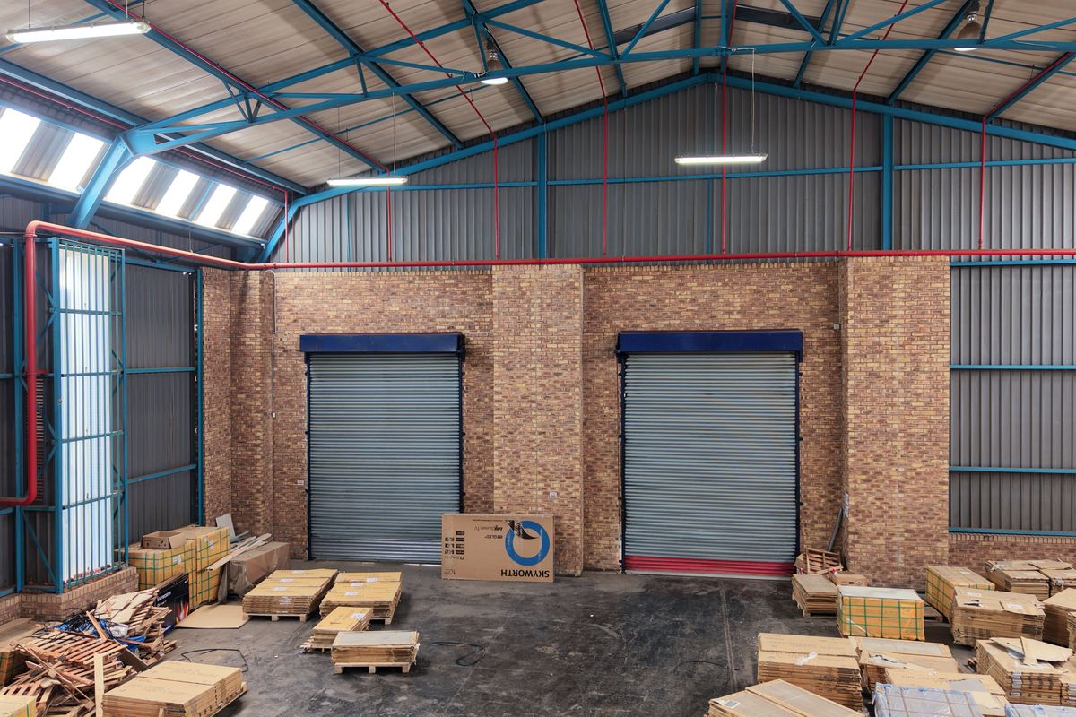 Spacious industrial warehouse unit featuring high ceilings with exposed steel trusses, brick walls, and two large roller shutter doors for easy access.
