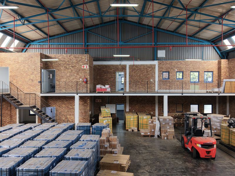 Spacious industrial warehouse featuring a mezzanine level, exposed steel roof trusses, brick walls, and high-clearance ceilings suitable for storage or manufacturing operations.