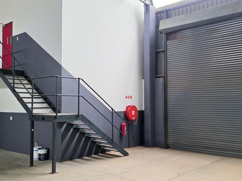 Industrial warehouse space featuring a large roller shutter door, concrete flooring, and a metal staircase leading to an elevated mezzanine level.