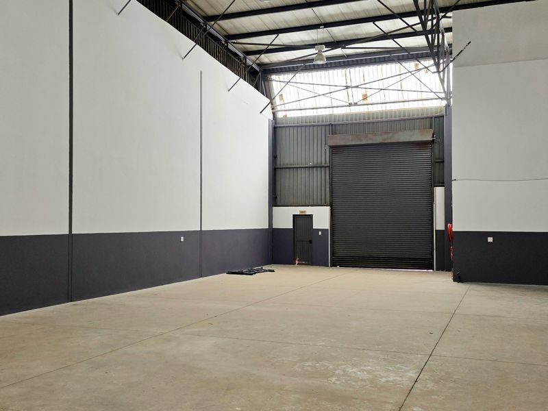 Spacious industrial warehouse unit featuring high ceilings, exposed steel trusses, concrete flooring, and a large roller shutter door for easy access.