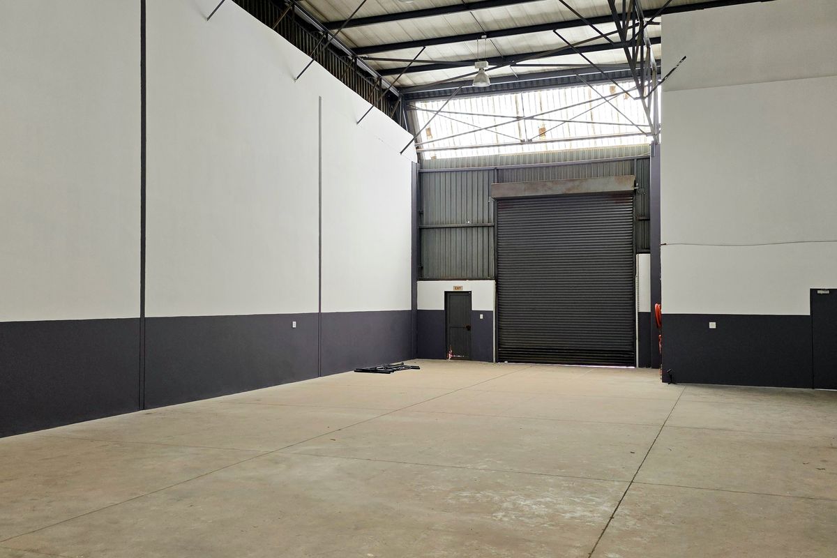 Spacious industrial warehouse unit featuring high ceilings, exposed steel trusses, concrete flooring, and a large roller shutter door for easy access.