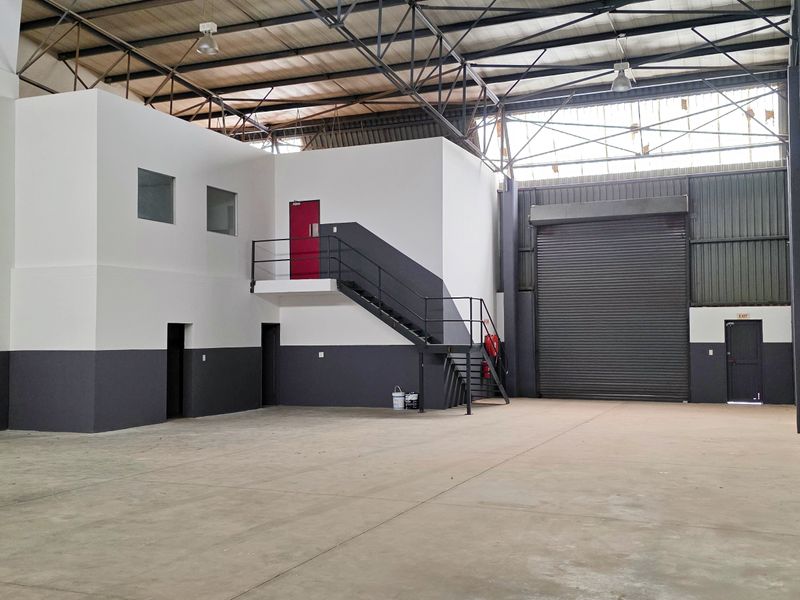 Spacious industrial warehouse unit featuring high ceilings with exposed steel trusses, a large roller shutter door, concrete flooring, and an integrated mezzanine office area.