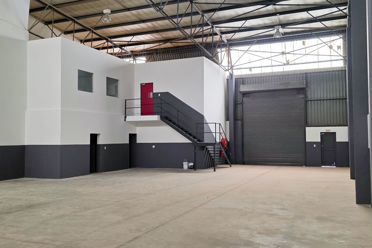 Spacious industrial warehouse unit featuring high ceilings with exposed steel trusses, a large roller shutter door, concrete flooring, and an integrated mezzanine office area.