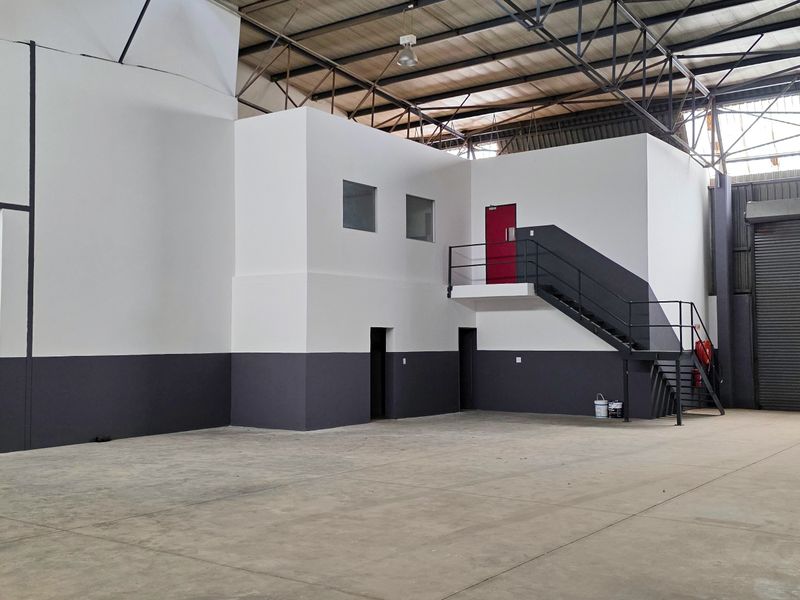 Spacious industrial warehouse unit featuring high ceilings with exposed steel trusses, concrete flooring, a roller shutter door, and a mezzanine level with staircase access.