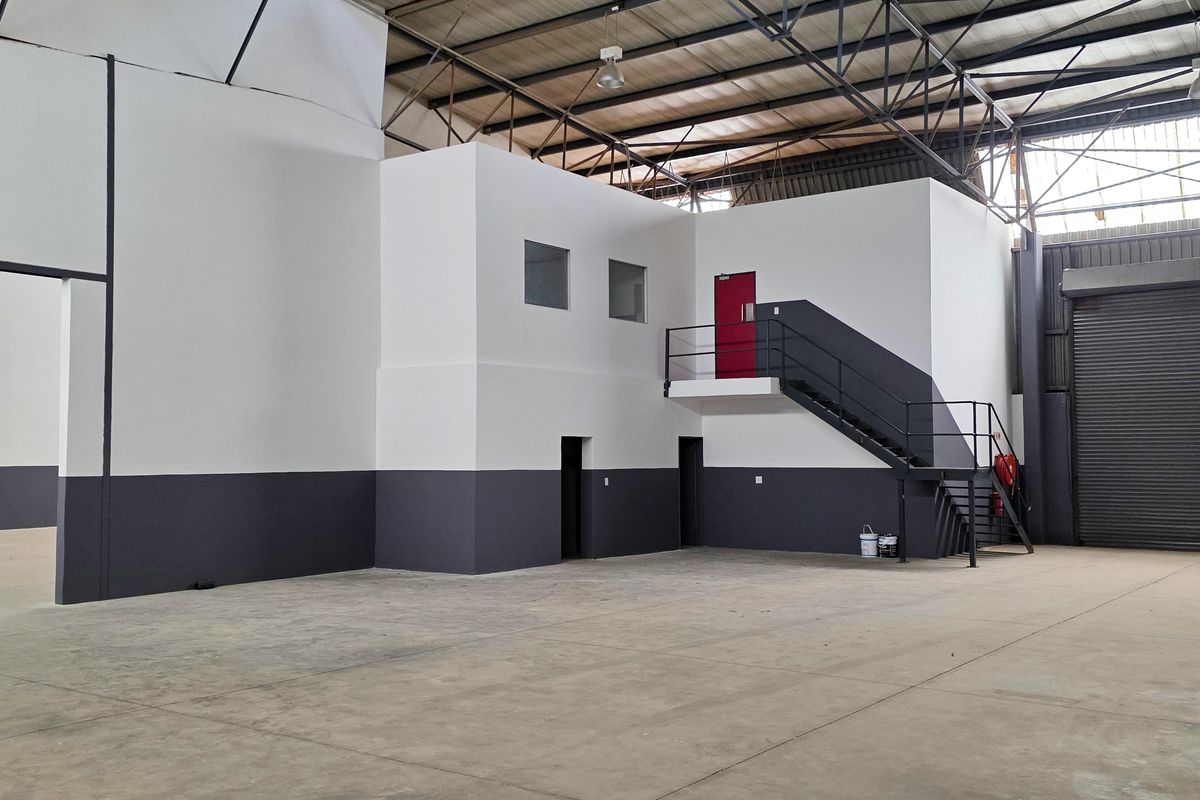 Spacious industrial warehouse unit featuring high ceilings with exposed steel trusses, concrete flooring, a roller shutter door, and a mezzanine level with staircase access.