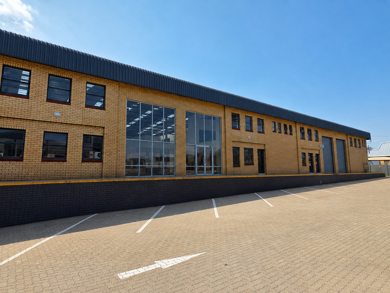 A modern industrial warehouse facility featuring a brick facade, large glass entrance, roller shutter doors, and an expansive paved parking and loading area.