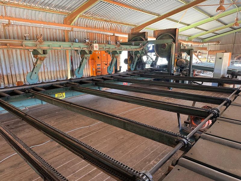 Industrial sawmill facility featuring heavy-duty timber processing machinery, conveyor systems, and a large-span corrugated metal structure.