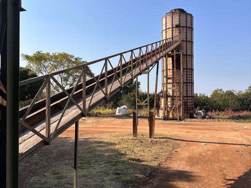 Agricultural utility area featuring a tall grain silo connected to a metal conveyor belt structure on a dirt farm yard.