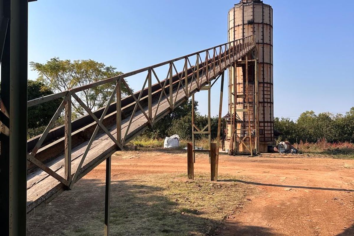Agricultural utility area featuring a tall grain silo connected to a metal conveyor belt structure on a dirt farm yard.