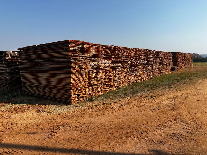 Large stacks of processed timber stored outdoors on a farm property, situated on a dirt access track.