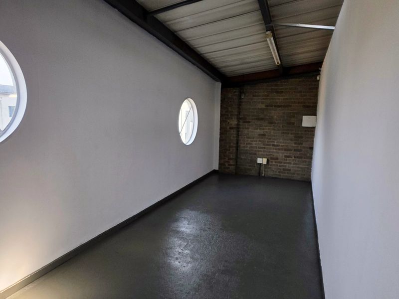 An empty industrial utility space featuring exposed brickwork, circular windows, and an exposed steel roof structure with durable epoxy flooring.