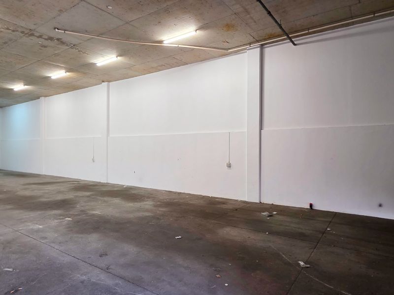 Spacious industrial utility area featuring high concrete ceilings, fluorescent lighting, and a large roller shutter door, suitable for storage or workshop use.