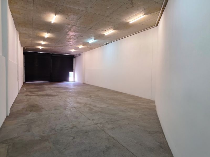 A spacious, empty industrial utility area featuring an exposed concrete ceiling, fluorescent lighting, and a large roller shutter door for easy access.