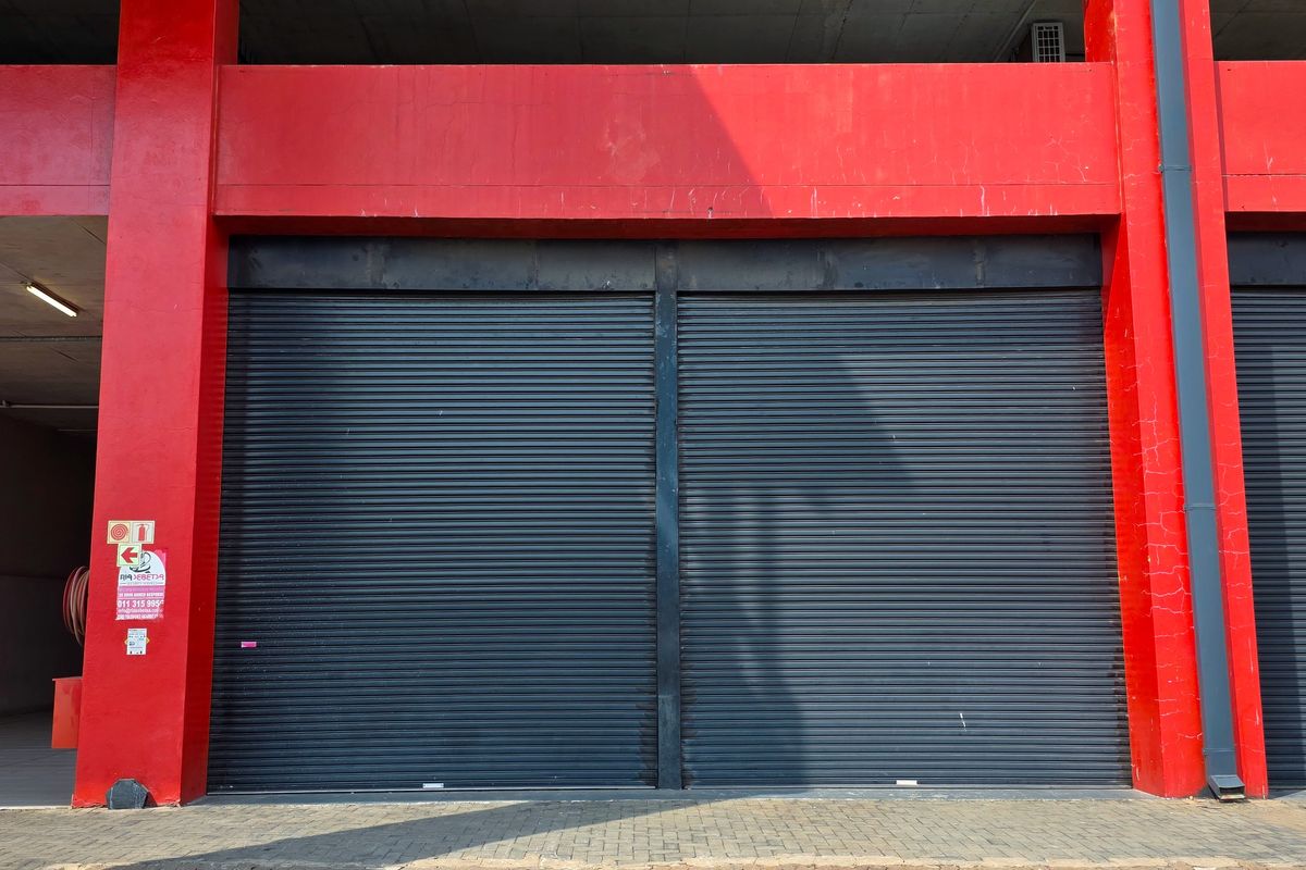 Industrial property frontage featuring secure roller shutter doors, red structural steel beams, and a paved driveway area.