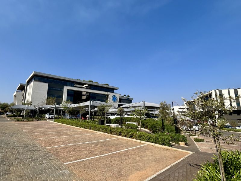 A modern multi-story office complex featuring extensive paved parking, shade cloth carports, and well-maintained landscaped garden areas.