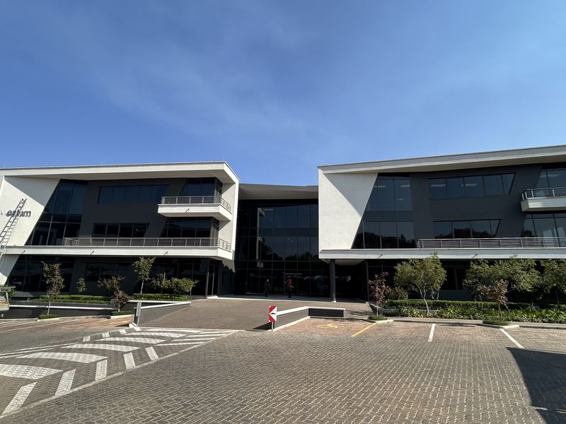 Modern multi-story office building featuring a glass facade, integrated balconies with metal railings, and a paved parking area.