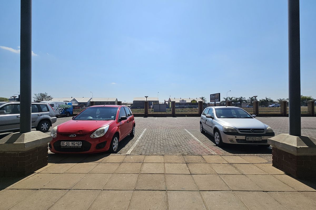 A paved outdoor parking area featuring brick perimeter walls and metal fencing, viewed from a covered entrance area.