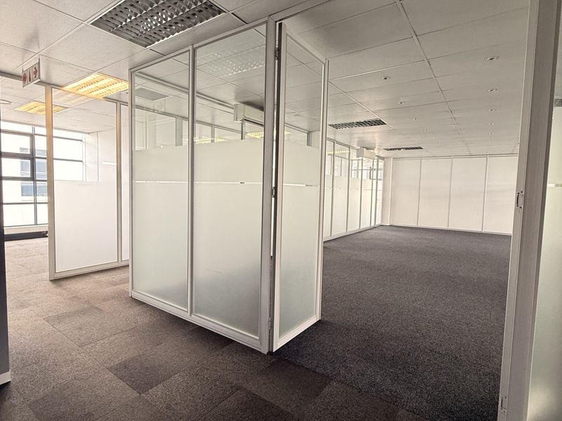 A spacious, vacant office area featuring glass partition walls, suspended ceilings with recessed lighting, and durable carpet tile flooring.
