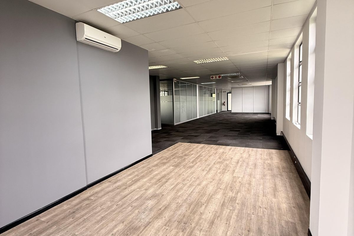 Spacious open-plan office area featuring a mix of wood-look vinyl and carpeted flooring, suspended ceilings, and glass partitions.
