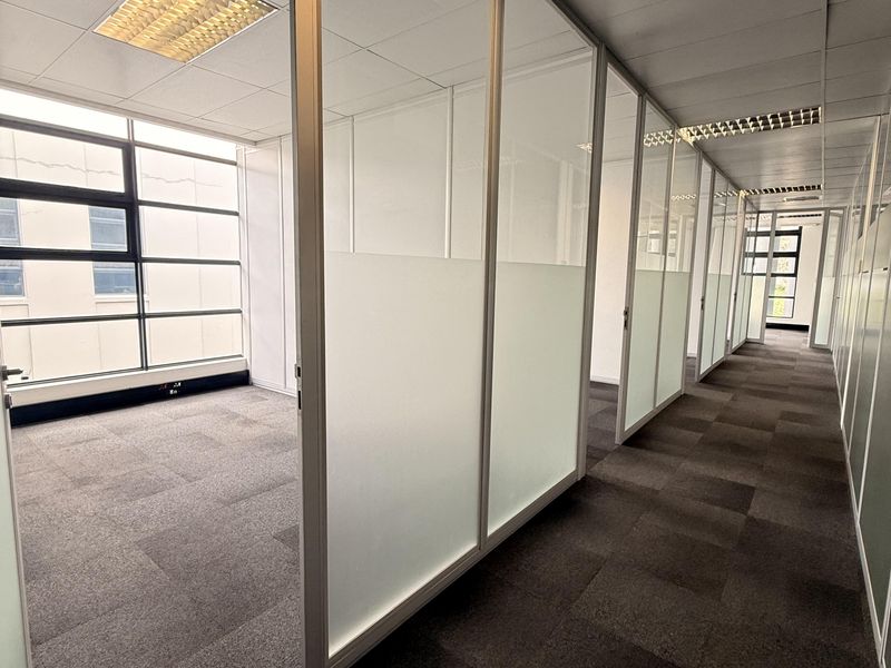 A bright, modern office space featuring glass partition walls, carpeted flooring, and large windows providing ample natural light.