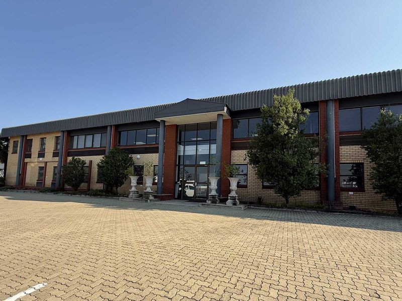 Front elevation of a commercial industrial building featuring a brick facade, metal roofing, and a paved parking area at the entrance.