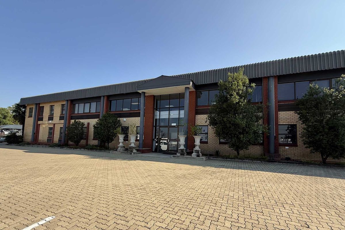 Front elevation of a commercial industrial building featuring a brick facade, metal roofing, and a paved parking area at the entrance.